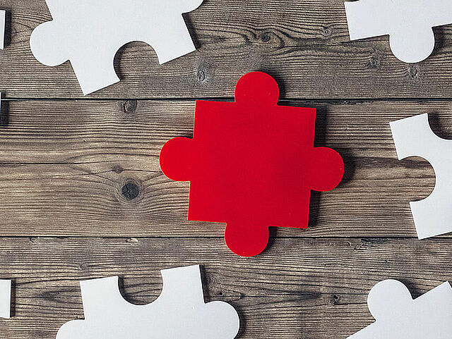 Mehrere Hände halten auf einem Holzuntergrund weiße Puzzleteile zusammen, in der Mitte liegt ein rotes.
