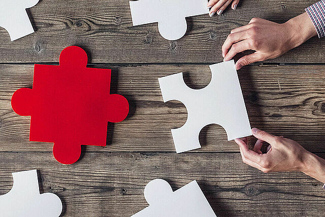 Mehrere Hände halten auf einem Holzuntergrund weiße Puzzleteile zusammen, in der Mitte liegt ein rotes.