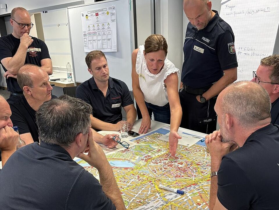 Planübung MANV Mehrere Personen, teilweise in Dienstkleidung der Polizei und Feuerwehr, sitzen und stehen um einen Tisch herum, auf dem ein Stadtplan liegt.