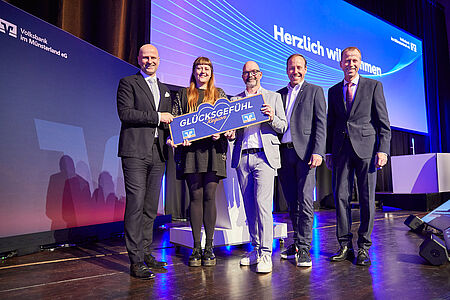 Fünf Personen stehen auf einer Bühne und lächeln in die Kamera. Zwei von ihnen halten gemeinsam ein großes, blaues Schild in Herzform mit der Aufschrift „GLÜCKSGEFÜHL Region“ sowie zwei Logos der Volksbank. Im Hintergrund ist ein großes Display mit der Aufschrift „Herzlich willkommen“ sowie dem Logo der Volksbank im Münsterland eG zu sehen. Die Personen sind festlich gekleidet.
