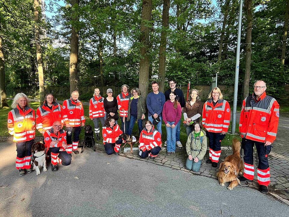 Gruppe von Menschen, teilweise in Einsatzkleidung, und Hunden vor einem Wald
