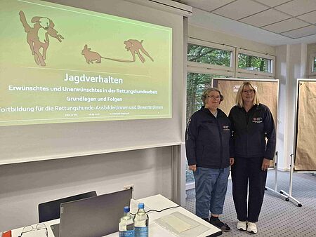 Zwei Frauen stehen neben einer Präsentation zum Thema Jagdverhalten von Hunden in der Rettungshundeeinheit