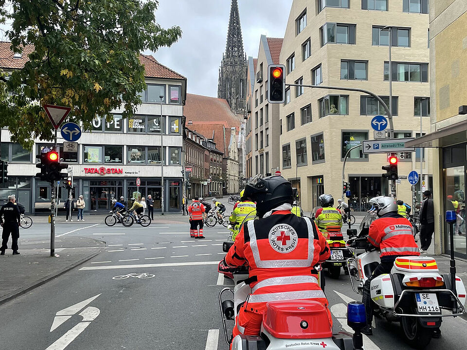 Einsatzkräfte auf Motorrädern auf einer Kreuzung in der Innenstadt, im Hintergrund Fahrradfahrer*innen und ein Kirchturm