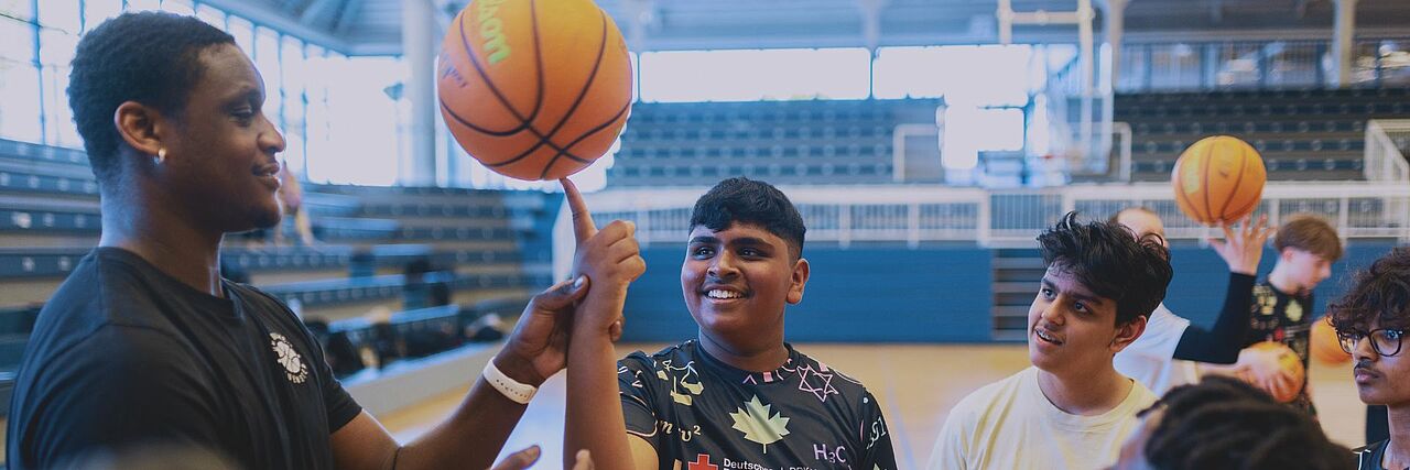 In einer Sporthalle stehen mehrere Jugendliche mit Basketbällen um einen Mann herum, der ihnen einen Trick mit dem Ball zeigt