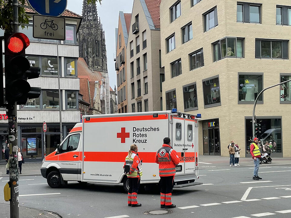 Zwei Einsatzkräfte des DRK stehen neben einem Krankenwagen an einer Kreuzung, moderne Gebäude und ein Kirchturm im Hintergrund.