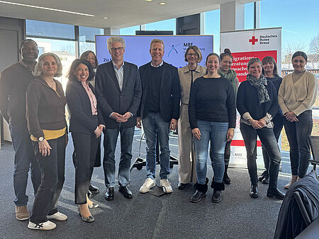 Gruppenbild Eine Gruppe von zehn Menschen, die in einem modernen Raum zusammenstehen und lächeln. Im Hintergrund ist ein Banner des Deutschen Roten Kreuzes zu sehen.