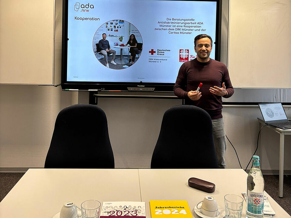 Ein Mann steht vor einer Präsentation und hinter einem Tisch, auf dem Informationsmaterialen liegen.