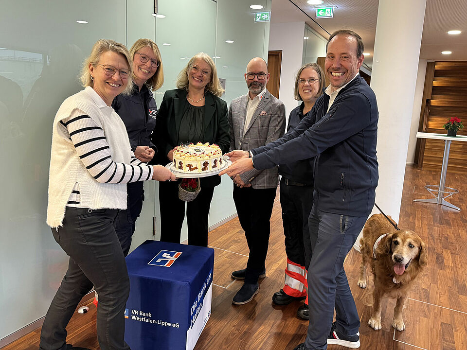 Sechs Personen stehen in einem Halbkreis und halten einen Kuchen in die Kamera