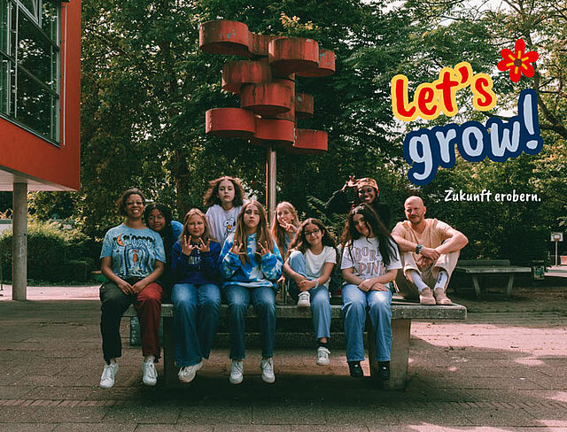 Eine Gruppe Mädchen sitzt und steht locker auf und um eine Betonbank auf einem Schulhof. Im Hintergrund ist ein moderner Bau und ein markantes rotes Kunstobjekt zu sehen. Der Slogan „Let’s grow! Zukunft erobern.“ ist grafisch eingeblendet.