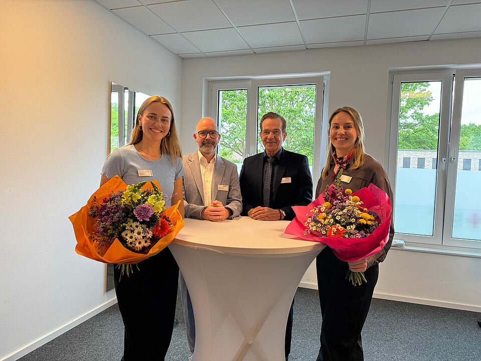 Vier Personen stehen um einen Stehtisch herum, die beiden Frauen außen halten einen Blumenstrauß in der Hand. Im Hintergrund ist ein moderner Raum mit Fensterfront zu sehen.