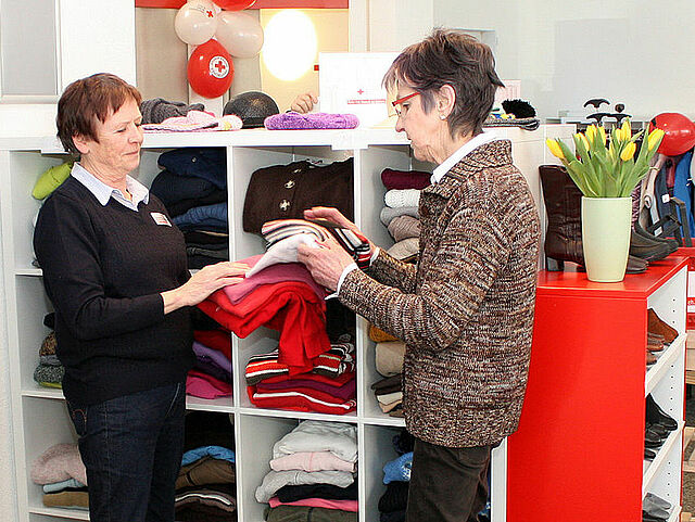 Zwei Personen im Gespräch in einem Bekleidungsgeschäft. Zwischen Regalen mit ordentlich gefalteter Kleidung und einem farbenfrohen Verkaufsbereich mit Tulpen und Ballons