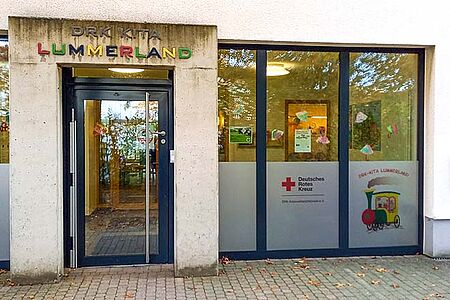 Lummerland Eingang eines Gebäudes mit dem Schild ‚DRK KITA LUMMERLAND‘ über der Tür. Der Schriftzug ‚LUMMERLAND‘ ist in bunten Buchstaben gestaltet. Links und rechts der Tür befinden sich große Glasfenster mit Dekorationen und Plakaten. Auf einem Fenster ist das Logo des Deutschen Roten Kreuzes sowie eine Illustration eines Zuges mit dem Schriftzug ‚DRK-KITA LUMMERLAND‘ zu sehen