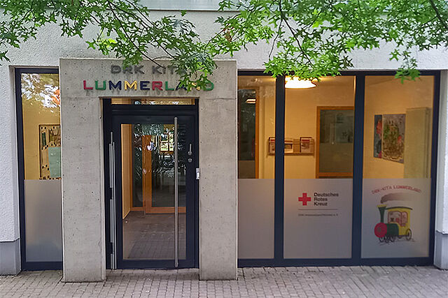 Eingang eines Gebäudes mit dem Schild ‚DRK KITA LUMMERLAND‘ über der Tür. Der Schriftzug ‚LUMMERLAND‘ ist in bunten Buchstaben gestaltet. Links und rechts der Tür befinden sich große Glasfenster mit Dekorationen und Plakaten. Auf einem Fenster ist das Logo des Deutschen Roten Kreuzes sowie eine Illustration eines Zuges mit dem Schriftzug ‚DRK-KITA LUMMERLAND‘ zu sehen