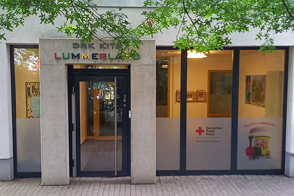 Lummerland Eingang eines Gebäudes mit dem Schild ‚DRK KITA LUMMERLAND‘ über der Tür. Der Schriftzug ‚LUMMERLAND‘ ist in bunten Buchstaben gestaltet. Links und rechts der Tür befinden sich große Glasfenster mit Dekorationen und Plakaten. Auf einem Fenster ist das Logo des Deutschen Roten Kreuzes sowie eine Illustration eines Zuges mit dem Schriftzug ‚DRK-KITA LUMMERLAND‘ zu sehen
