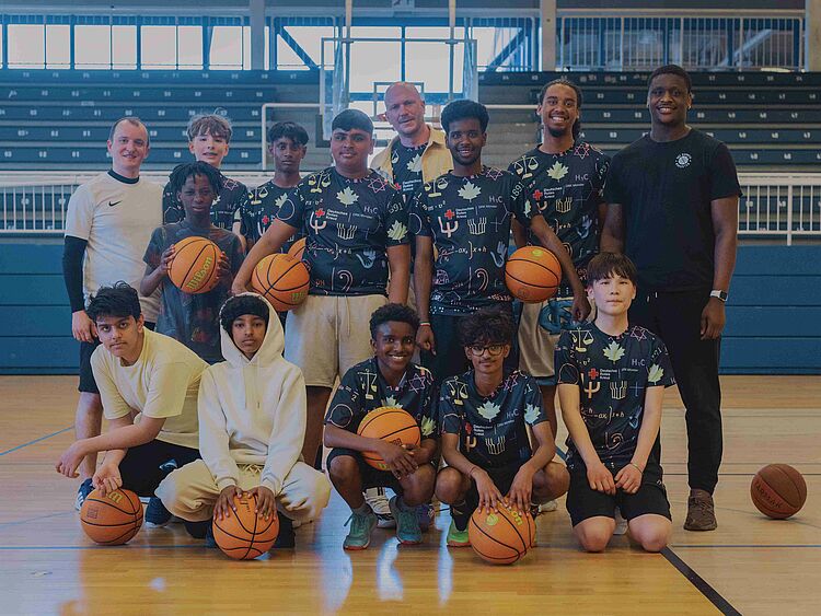 Gruppenbild von Jugendlichen und Erwachsenen in einer Sporthalle. Sie tragen Trikots mit der Aufschrift "Hood Hoopers" und halten Basketbälle.