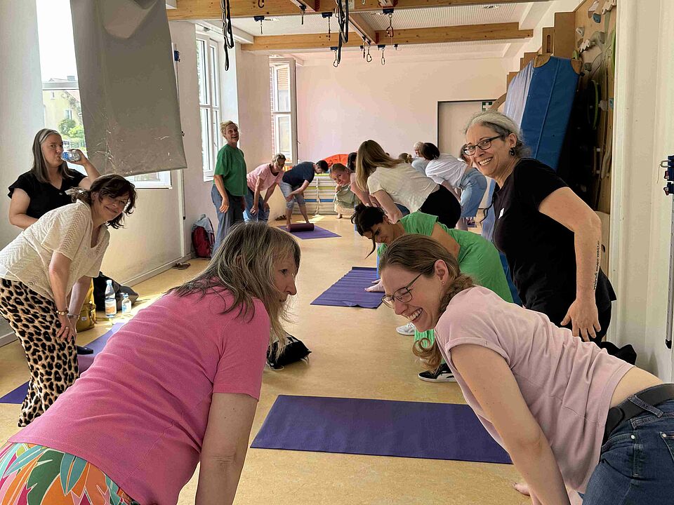 Eine Turnhalle mit Menschen auf Matten, die Dehnübungen machen und teilweise in die Kamera lachen