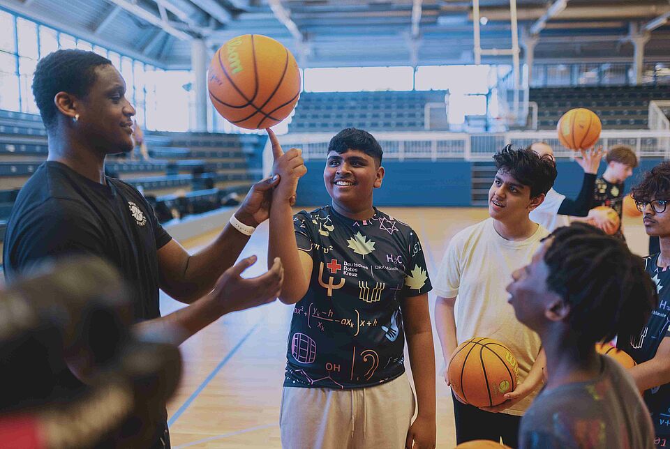 Hood Hoopers In einer Sporthalle stehen mehrere Jugendliche mit Basketbällen um einen Mann herum, der ihnen einen Trick mit dem Ball zeigt
