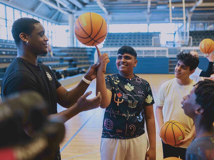 In einer Sporthalle stehen mehrere Jugendliche mit Basketbällen um einen Mann herum, der ihnen einen Trick mit dem Ball zeigt