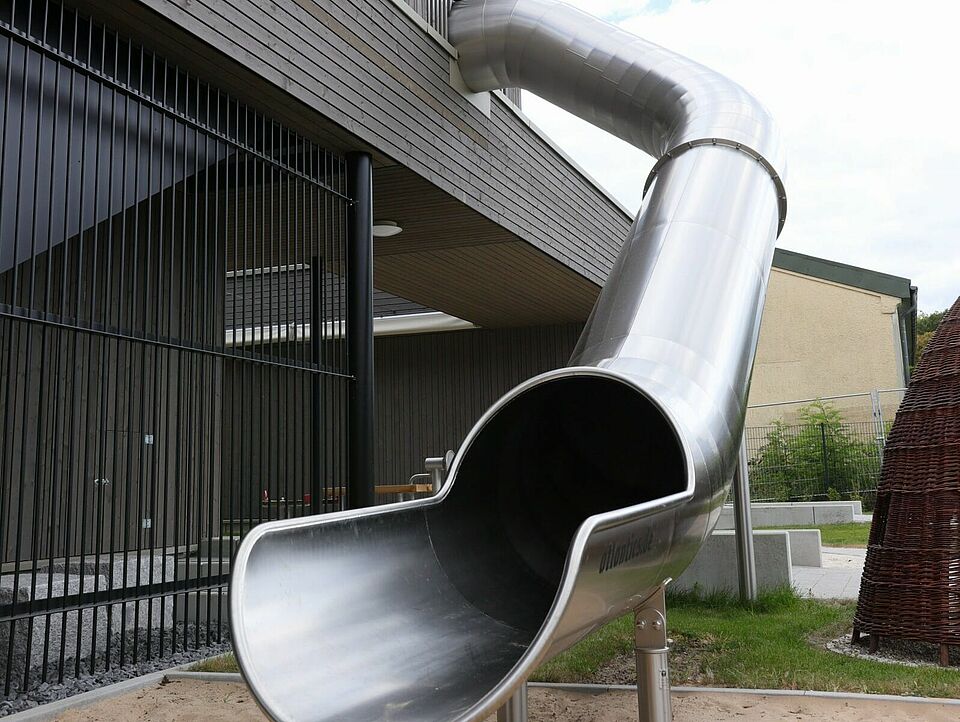 Eine silberne Rutsche führt aus dem ersten Stock eines modernen Gebäudes runter auf eine Sandfläche