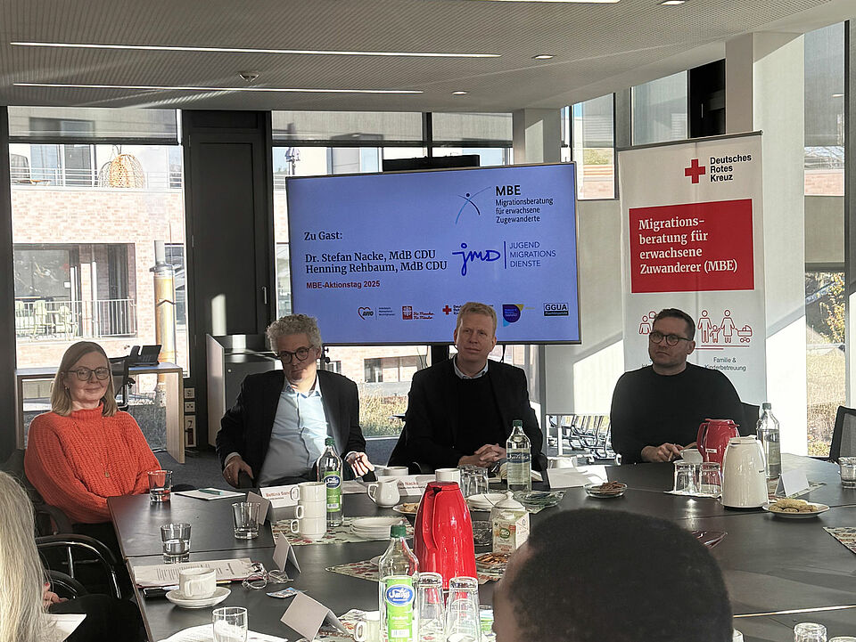 Vier Personen, die an einem Tisch sitzen. Auf der linken Seite ist eine Frau mit rotem Pullover zu sehen, daneben zwei Männer, einer mit grauen Haaren und Brille, der andere mit einer schwarzen Jacke. Der vierte Mann, rechts im Bild, trägt ein schwarzes Shirt. Im Hintergrund sind ein Bildschirm mit einer Präsentation und ein Banner des Deutschen Roten Kreuzes zu erkennen