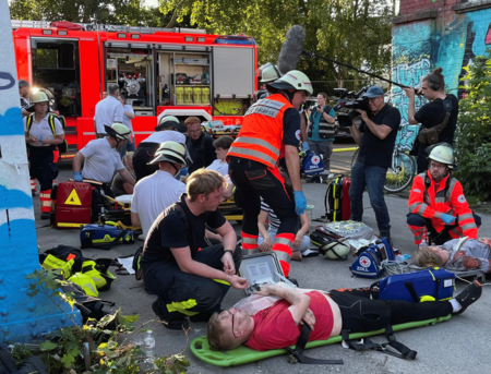 Mehrere Verletztende liegen auf Tragen auf dem Boden und werden von Einsatzkräften versorgt. Im Hintergrund ist ein Feuerwehreinsatzfahrzeug zu sehen.