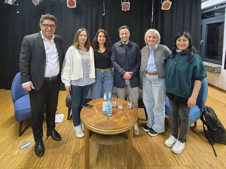 Gruppenbild von sechs Personen in einem großen Raum. Vor ihnen steht ein kleiner Holztisch mit Getränken, im Hintergrund sind schwarze Vorhänge und Scheinwerfer zu sehen.
