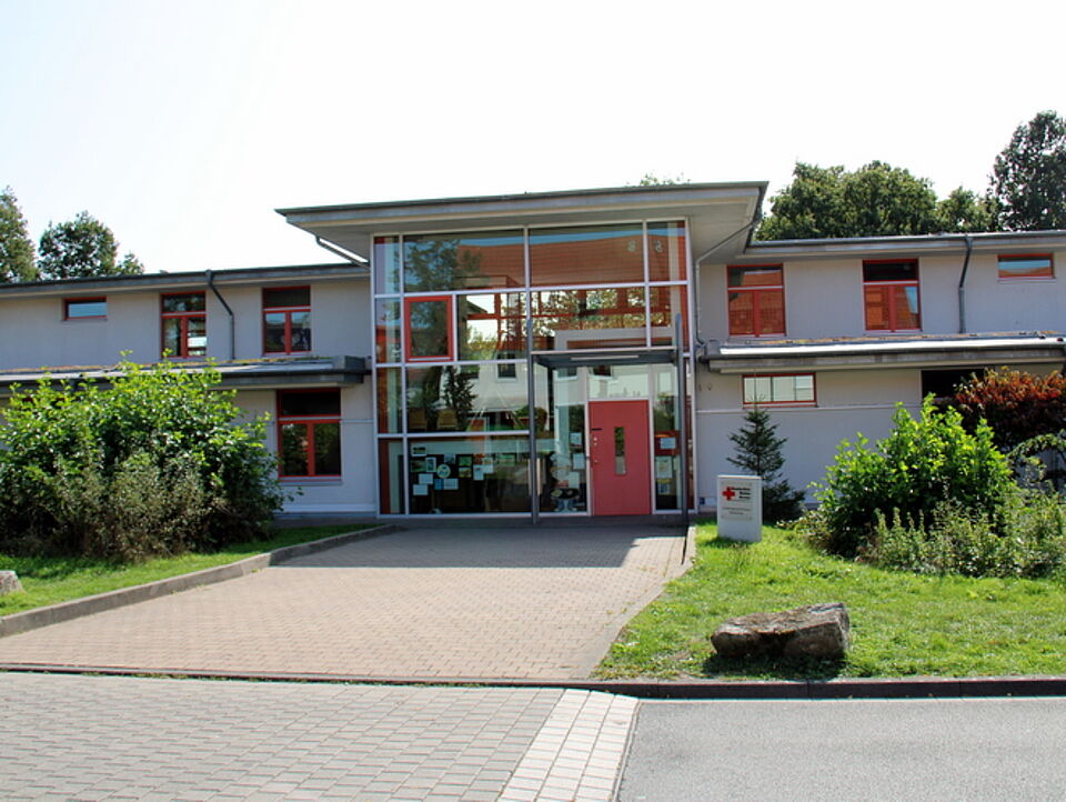 Ein modernes, zweistöckiges Gebäude mit großen Fenstern und einer roten Eingangstür. Ein gepflasterter Weg führt durch grüne Büsche und Bäume zum Eingang.