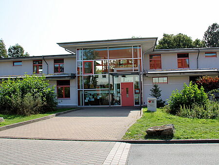 Ein modernes, zweistöckiges Gebäude mit großen Fenstern und einer roten Eingangstür. Ein gepflasterter Weg führt durch grüne Büsche und Bäume zum Eingang.