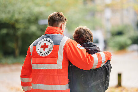 Es sind zwei Personen von hinten zu sehen. Eine Person trägt eine Einsatzjacke vom DRK und hat einen Arm um die andere Person in Zivil.