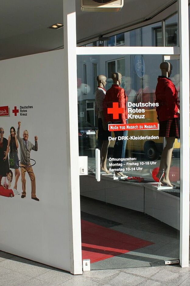 Eingang eines DRK-Kleiderladens mit Glastür. Auf der Tür steht: ‚Deutsches Rotes Kreuz – Kleidung für Mensch zu Mensch! Der DRK-Kleiderladen. Öffnungszeiten: Montag – Freitag 10–18 Uhr, Samstag 10–14 Uhr.‘ An der Wand neben der Tür hängt ein Plakat mit mehreren Personen und dem Text: ‚Kleidung für Mensch zu Mensch. Deutsches Rotes Kreuz