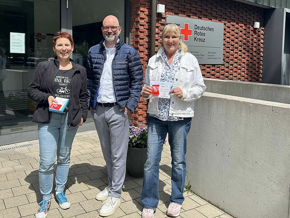 Vor dem Gebäude des Deutschen Roten Kreuzes in Münster stehen drei Personen lächelnd für ein Foto. Zwei Frauen halten die Preise in der Hand