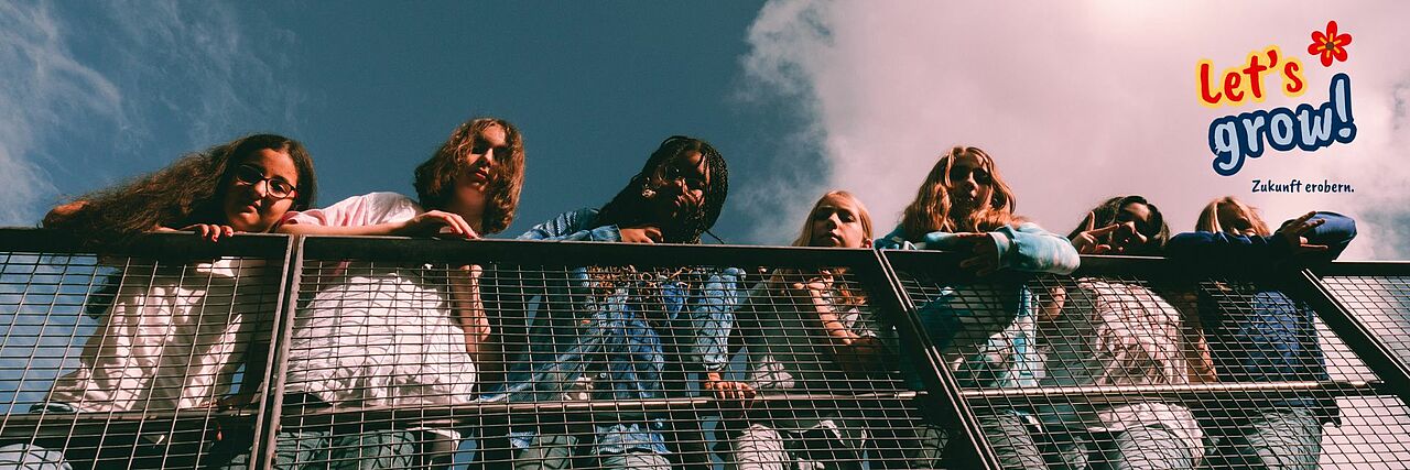 Eine Gruppe Mädchen lehnt lässig an einem hohen Metallgeländer, fotografiert aus der Froschperspektive mit blauem Himmel im Hintergrund.Der Schriftzug „Let’s grow! Zukunft erobern.“ ist eingeblendet.