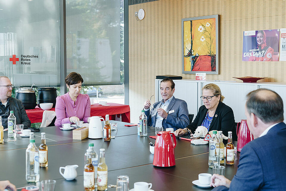 Mehrere Personen sitzen um einen Konferenztisch mit Getränken und Thermoskannen, im Hintergrund ist das DRK-Logo sichtbar.