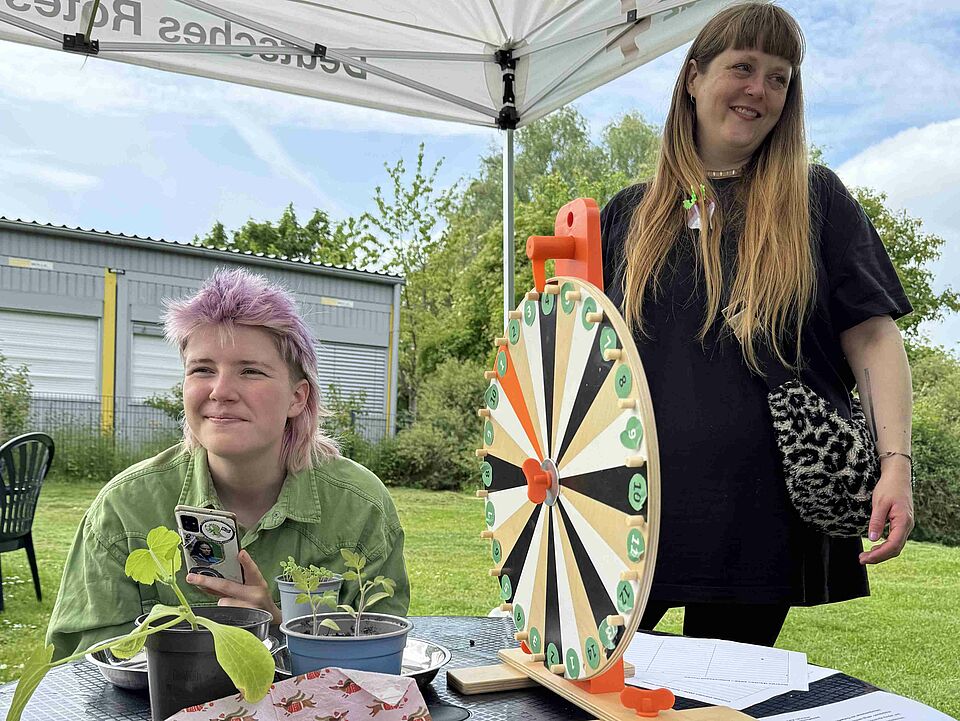Coerde Garden Glücksrad Zwei personen unter einem Pavillion und an einem Tisch, auf dem ein Glücksrad und Pflanzen stehen