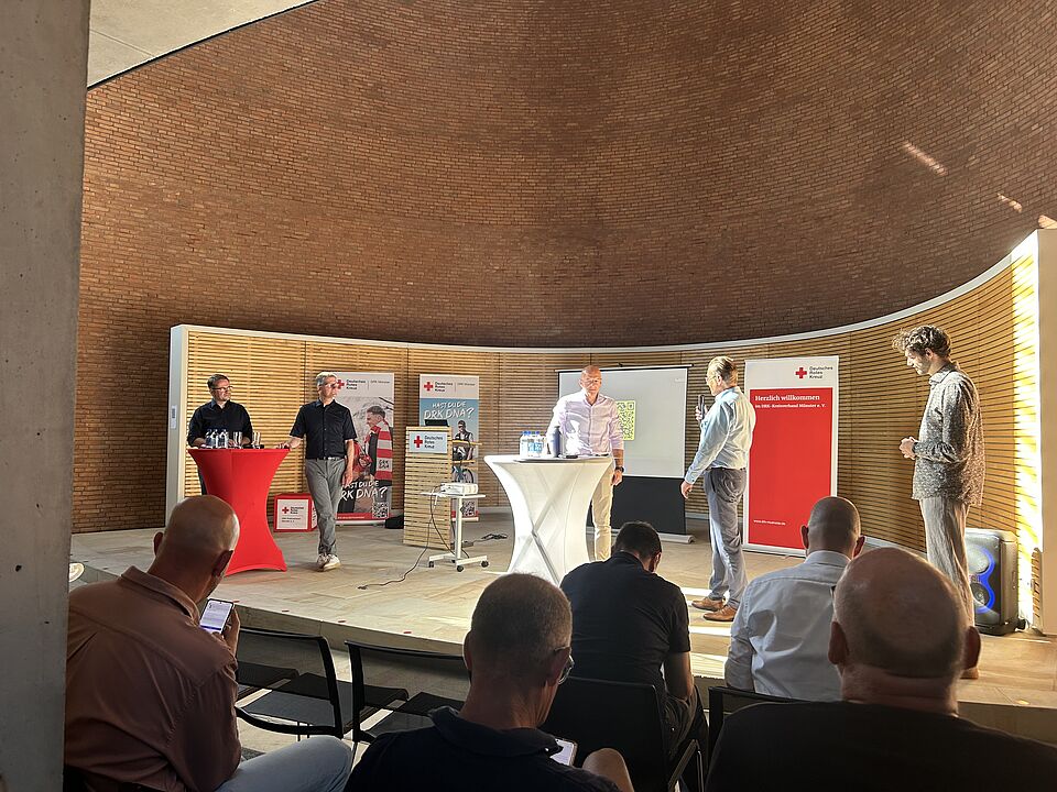 Fünf Personen stehen auf einer Bühne vor einem sitzenden Publikum. Im Hintergrund sind Holzwände und Banner mit dem Rotkreuz-Logo zu sehen