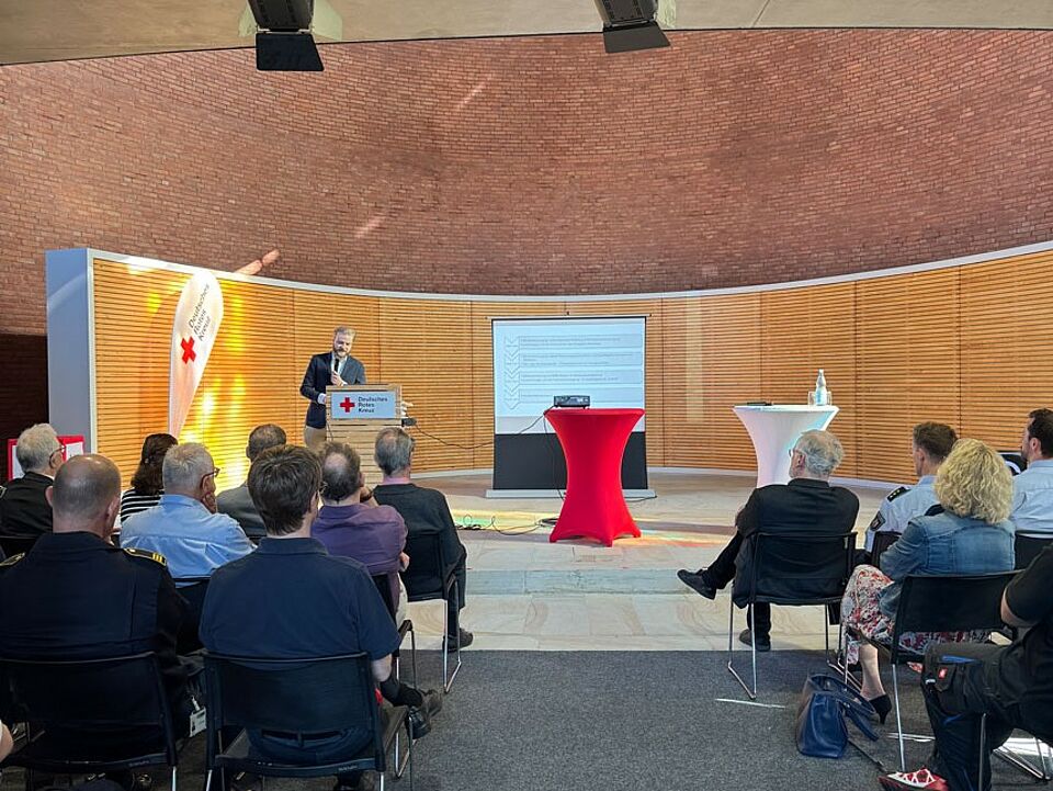 Eine kleine Bühne in einem Backsteingebäude mit einem kleinen Publikum, zwei Stehtischen, einer Leinwand und einer Person an einem Rednerpult.