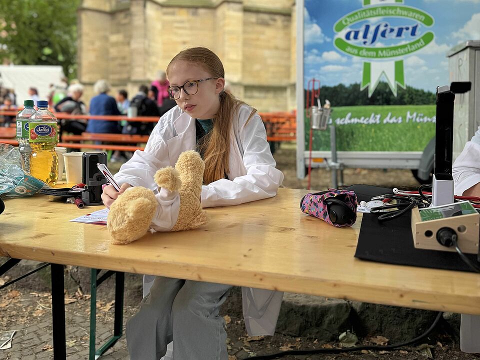 Ein Kind sitzen an einem Tisch unter einem Zelt. Auf dem Tisch liegt ein Teddy und mehrere Utensilien.. Das Kind trägt ein türkises T-Shirt und einen weißen Kittel und hält einen Stift in der Hand.