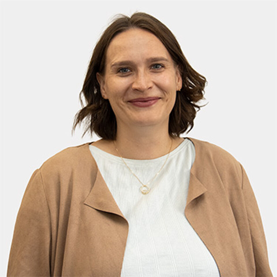 Frau mit braunen Haaren, blauer Bluse und schwarzem Blazer vor weißem Hintergrund