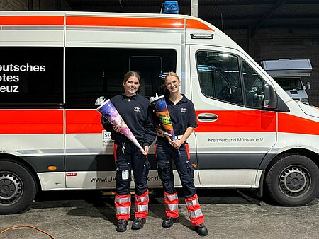 Zwei Personen stehen in Einsatzkleidung mit Schultüten vor einem Krankentransportwagen des DRK Münsters. Im Hintergrund ist eine Fahrzeughalle zu sehen.