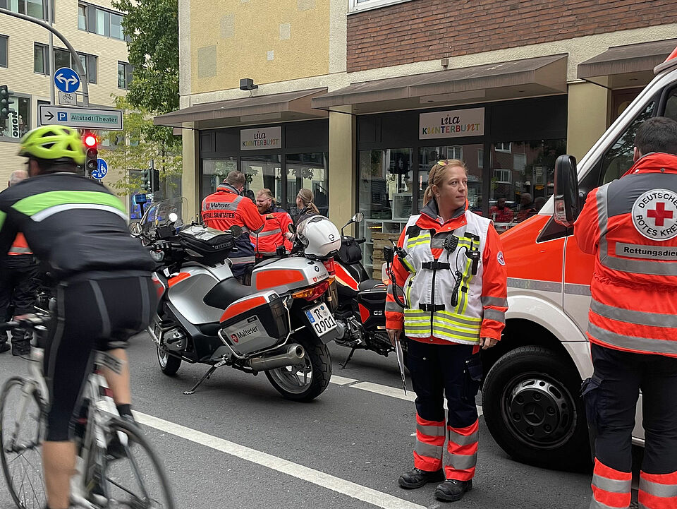 Rettungskräfte stehen neben Motorrädern und einem Rettungsmittel am Straßenrand, ein Radfahrer fährt vorbei.