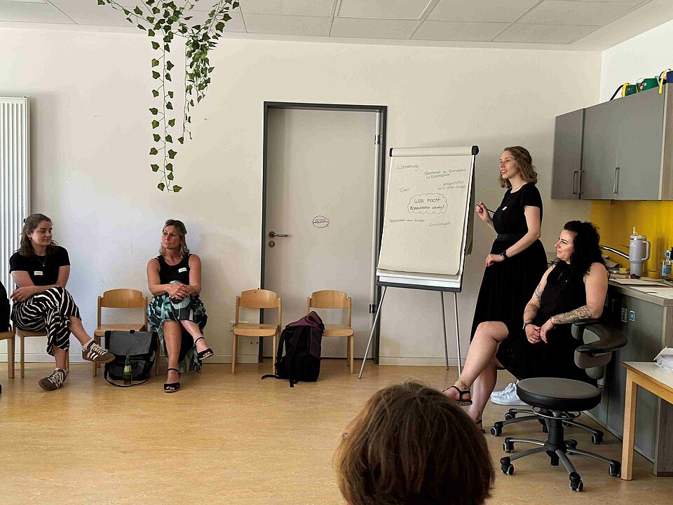 Zwei Personen neben einer Flipchart, eine davon schreibt auf der Flipchart in einem Raum mit aufgestellten Stühlen und zwei sichtbaren Personen