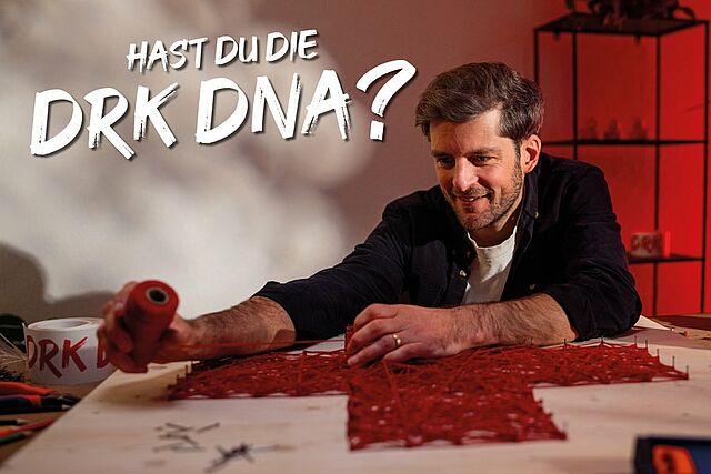 Eine Person sitzt an einem Tisch und arbeitet mit rotem Faden an einem Kunstprojekt in Rotkreuzform. Text im Bild: „Hast du die DRK DNA?“