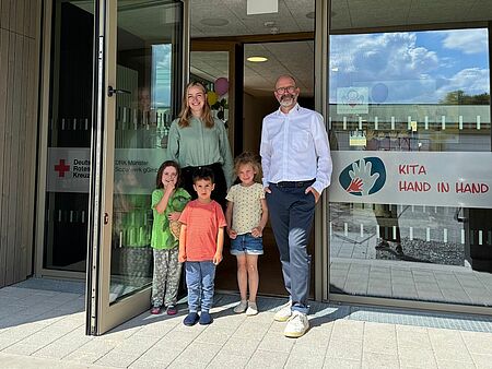 Zwei Personen mit drei Kindern stehen in einer Tür von einem neuen Gebäude. Auf den Glasfronten ist das Rotkreuzlogo und das Logo der Kita Hand in Hand zu sehen.
