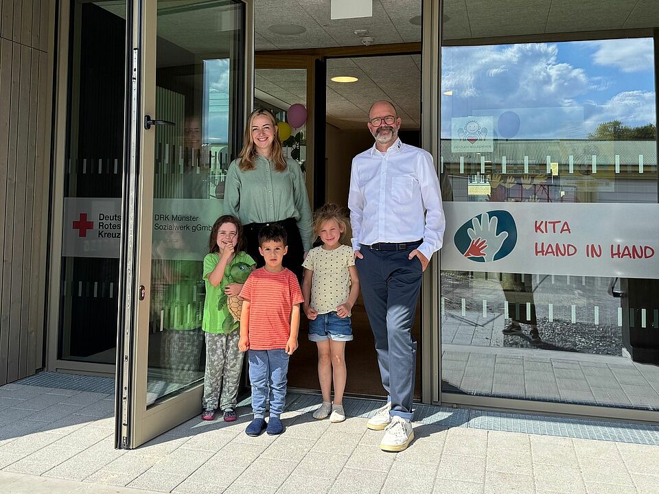 Zwei Personen mit drei Kindern stehen in einer Tür von einem neuen Gebäude. Auf den Glasfronten ist das Rotkreuzlogo und das Logo der Kita Hand in Hand zu sehen.