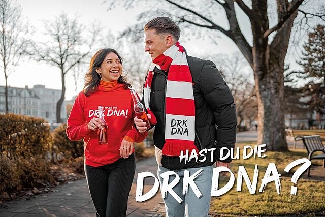 Zwei Personen gehen im Freien in einer parkähnlichen Umgebung und trinken rote Getränke. Eine trägt einen roten Pullover mit der Aufschrift „Ich habe die DRK DNA!“, die andere eine schwarze Jacke und einen rot-weiß gestreiften Schal mit „DRK DNA“. Unten rechts steht der Text: „Hast du die DRK DNA?“